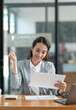 © crizzystudio - Asian businesswoman sitting happily in office holding detailed report paper analyzing data accuracy in financial matters management concept.