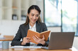© crizzystudio - Beautiful Asian businesswoman working on a laptop and having fun at work and reading review book documenting detailed financial information earning peace of mind.