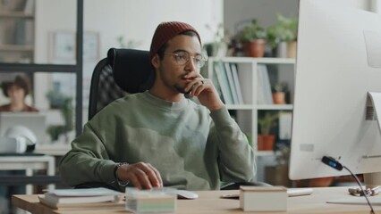 Medium shot of young programmer sitting at his workplace in front of computer monitor and working with program