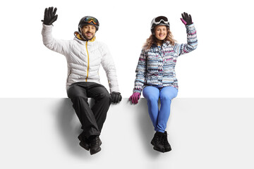 Sticker - Young couple wearing winter skiing clothes sitting on a blank panel and waving