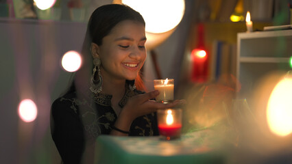 Smiling pretty female wearing big earrings sitting and hold burning glass candles in hand closer to face on the occasion of Diwali. Teenager enjoy Deepawali festival with Decorative surroundings