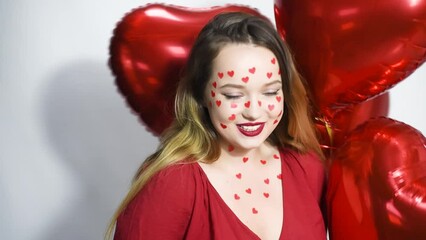 Wall Mural - A woman with heart stickers on her face. The girl on the background of red balloons in the shape of a heart. Love, feelings. The girl dances, laughs, air kisses.