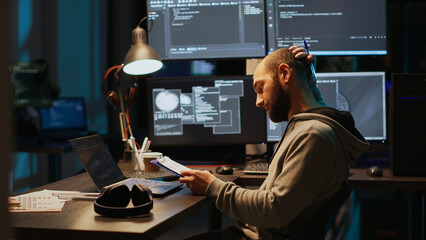 Male IT engineer analyzing web data on clipboard papers and screens, comparing information on files. Young man using programming language and coding new user interface at night.
