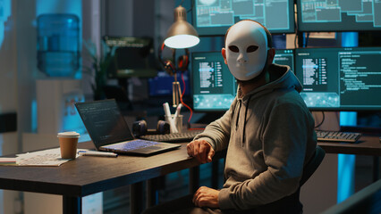 Masked scammer hacking computer network late at night, working on laptop and multiple monitors to steal IT information. Hacker with hidden identity breaking security server, cyber attack.