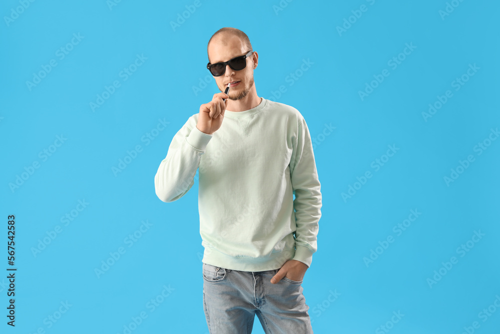 Young man in sunglasses with disposable electronic cigarette on blue background