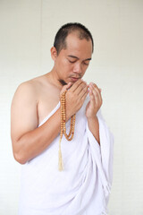 Wall Mural - Asian muslim man wearing white ihram clothes and praying while holding prayer beads