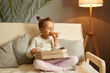 © sementsova321 - Indoor shot of cute funny child biting big slice pizza, sitting on sofa with crossed legs, enjoying tasty junk food, positive female kid with two bun hairstyle having dinner, eating fast food.