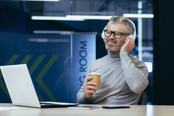 Mature businessman in headphones listening to audio books online podcasts and radio with music, man working inside office at laptop, boss smiling happy with achievement results and work.