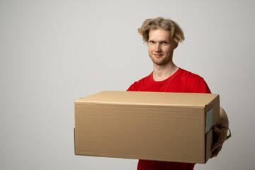 Wall Mural - Smiling young delivery man in red t-shirt standing with big parcel post box isolated over white background.