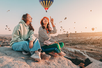 Wall Mural - Girl friends sitting on a clifftop viewpoint and admiring view of majestic flying hot air balloons in Cappadocia