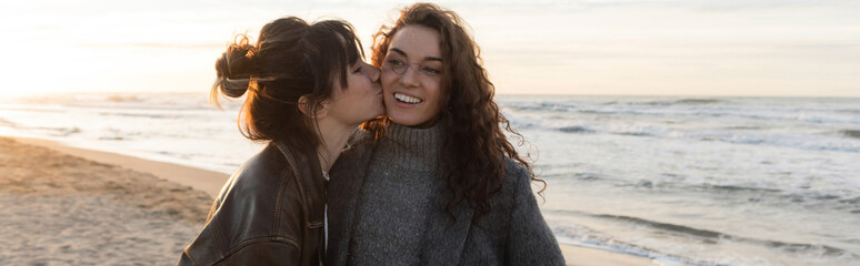 Wall Mural - Young woman kissing cheeks of cheerful friend on beach in Spain, banner.