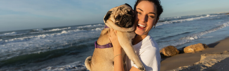 Wall Mural - cheerful young woman with curly hair holding pug dog on beach near sea in Barcelona, banner.