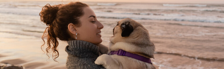 Wall Mural - Side view of cheerful young woman looking at pug dog near sea in Spain, banner.