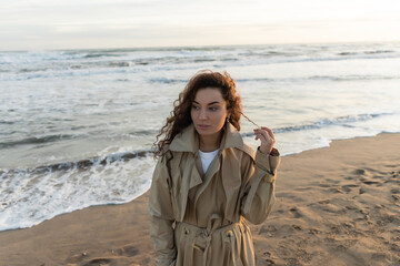 Wall Mural - Young woman in trench coat touching curly hair on beach.