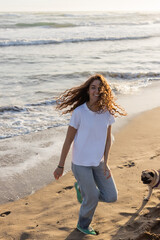 Sticker - cheerful young woman running with pug dog on sandy beach during sunset.