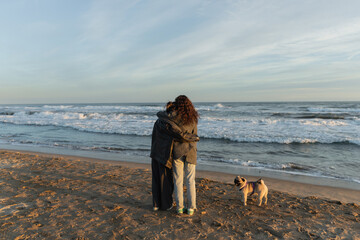 Wall Mural - Back view of young women hugging near pug dog on beach in Spain.