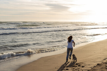 back view of woman running with pug dog on sandy beach during sunset.