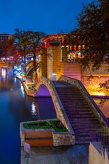 Wall Mural - Vibrant night landscape with warm colors of boardwalk, stone bridge, and water reflections on San Antonio River Walk canal on a quiet winter night with empty footpath in Texas, USA