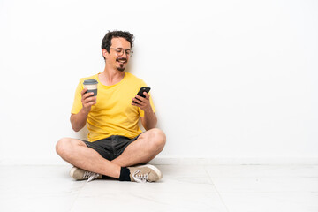 Wall Mural - Young caucasian man sitting on the floor isolated on white background holding coffee to take away and a mobile