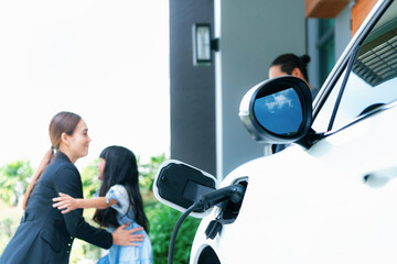 Focus closeup electric vehicle recharging battery from home electric charging station with blurred family in background. Renewable clean energy car for progressive eco awareness lifestyle concept.