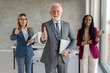 © Dorde - Confident gray-haired senior businessman wearing business suit holding laptop, smiling, looking at camera and showing thumbs up with two businesswoman a part of his team while standing in office.