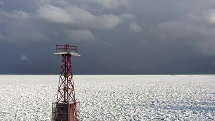 Wall Mural - Aerial flying up from a snow and ice covered Lake Michigan towards a pier with a steel red tower structure at the end of it and cloudy blue and gray sky above.