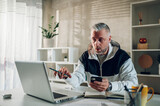 Fototapeta Na ścianę - Middle aged man using smartphone and a laptop while working in a home office