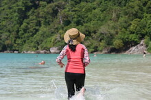 Woman Tourists Play In Sea Having Fun During Their Vacation. Stand With Her Back And Enters The Sea In Hat And Black-orange Slim Fit. Running Along The Shore In Foam Of Waves. Summer Vacation Concept.