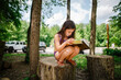 © Cavan Images - A little girl sits on tree stump reading large book in sunlight