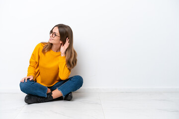 Wall Mural - Young caucasian woman sitting on the floor isolated on white background listening to something by putting hand on the ear