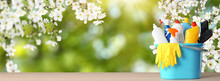 Spring Cleaning. Bucket With Detergents And Tools On Wooden Surface Under Blossoming Tree Against Blurred Green Background, Space For Text. Banner Design