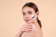 Beauty portrait of a beautiful half naked woman applying face cream isolated on studio background. Skin care product. Moisturizing creme on face. Perfect skin, morning routine.