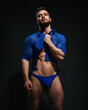 Sexy muscled male model in blue shirt and blue swimwear on black background. Bearded man taking his clothes up. Fashion guy in beachwear in studio. Male fitness model in stylish outlook.