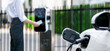 Focus electric vehicle recharging at public charging station with blurred progressive woman using credit card payment for electric refuel for her EV car for alternative eco-friendly car concept.