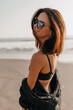 Spectacular lovely girl with short hair wearing back sunglasses and black shirt turn around to camera with happy smile on background of ocean shore