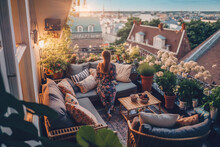 Girl Standing On A Big Balcony With Lots Of Flowers And Cosy Furniture Looking Into The Sunset Of The City, Generative Ai
