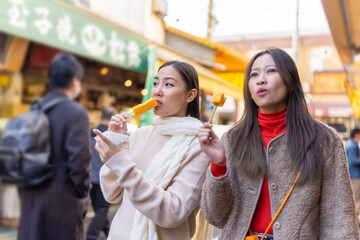 Asian woman friends eating street food sweet egg roll together while travel at fish market in Tokyo city, Japan. Attractive girl enjoy and fun outdoor lifestyle travel street market on autumn vacation