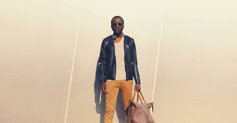 Wall Mural - Portrait of stylish young african man model posing wearing black rock leather jacket with bag on city street background