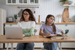 © DimaBerlin - Successful focused business woman doing work at laptop sits at kitchen table near daughter making homework. Thoughtful concentrated teenage girl studying on own for exams located near with busy mom