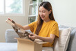 Happy excited, asian young woman, girl customer sitting on sofa at home, open and unpack cardboard box carton parcel after buy fashion bag, ordering present and shopping online, delivery service.