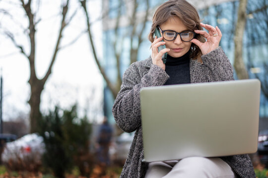 Mature adult caucasian businesswoman in glasses working on a laptop and talking on the phone while sitting on a bench against the backdrop of