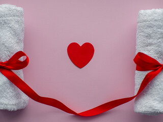 Flat lay with a tiny heart-shaped paper, two towels and space for text. Pink paper background with towels tied with a red bows. Valentines day spa, bath, self care and body care concept.