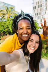 Vertical shot of two multiracial best female friends in casual summer clothes having fun together taking self portrait using phone outdoors.