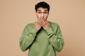 Wall Mural - Young surprised shocked confused man of African American ethnicity wear green sweatshirt cover mouth with hand isolated on plain pastel light beige background studio portrait People lifestyle concept