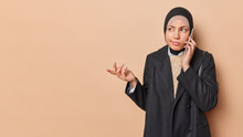 Waist Up Shot Of Questioned Arabian Woman Wears Black Hijab And Formal Jacket Calls Someone Has Hesitant Expression Looks Confused Isolated Over Brown Background Copy Space For Your Promotional Text