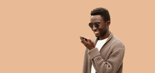 Wall Mural - Portrait of smiling young african man holding smartphone using voice command recorder, assistant or takes calling, looking at smartphone isolated on brown background