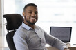 © fizkes - Cheerful confident young Black businessman sitting at office work table with laptop, investment, financial charts on screen, looking at camera with toothy smil. Corporate male portrait, headshot