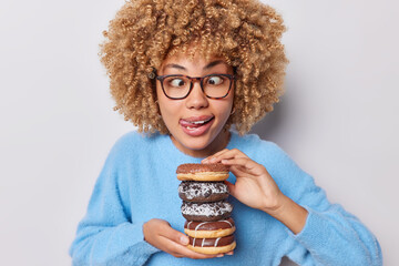 Wall Mural - Horizontal shot of funny curly haired woman foolishes around sticks out tongue crosses eyes wears transparent eyeglasses and blue jumper holds delicious sweet doughnuts poses against white background