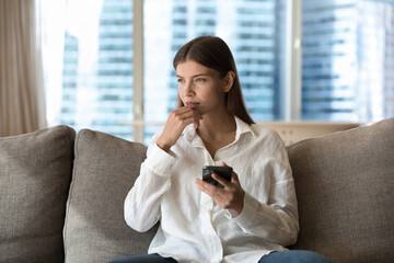 Wall Mural - Pensive young millennial woman lost in thoughts holding cellphone, looking away, thinking, dreaming, touching chin, making decision, typing, making phone call, sitting on home couch