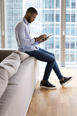 Wall Mural - Focused young entrepreneur man using tablet at home for Internet communication, working on job tasks, making payment with bank application, sitting on sofa against big window city view